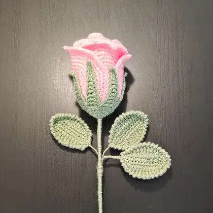 A crocheted bouquet with a large pink rose, red strawberries and green leaves wrapped in pink paper