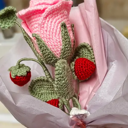 A crocheted bouquet with a large pink rose, red strawberries and green leaves wrapped in pink paper