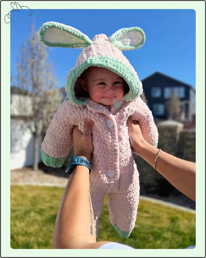 A baby wearing a pale pink crocheted bunny suit with mint trim and long bunny ears on the hood, held up outdoors