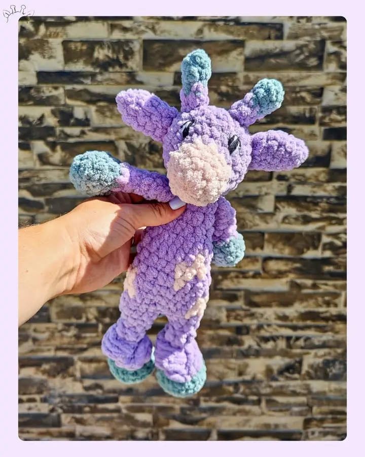 A purple crocheted giraffe with teal hooves and cream patches, held in one hand against a stone wall