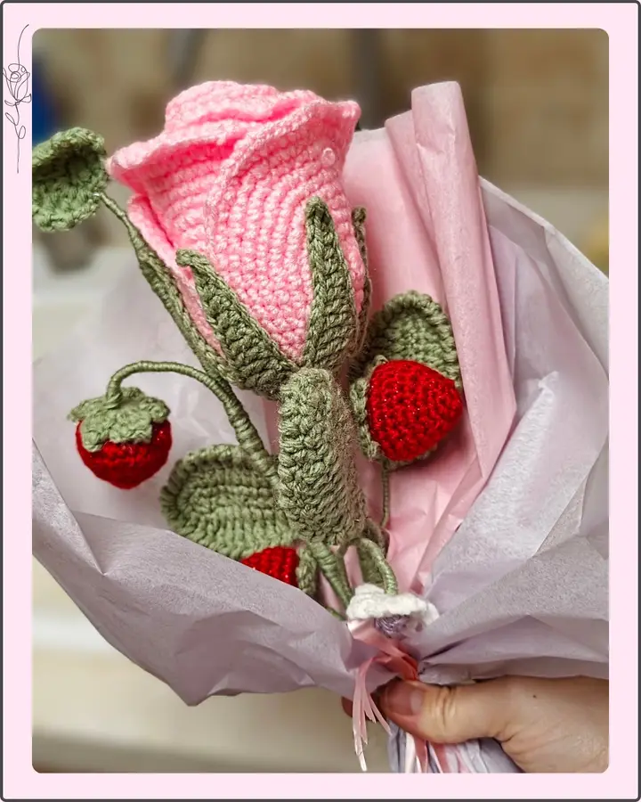 A crocheted bouquet with a large pink rose, red strawberries and green leaves wrapped in pink paper
